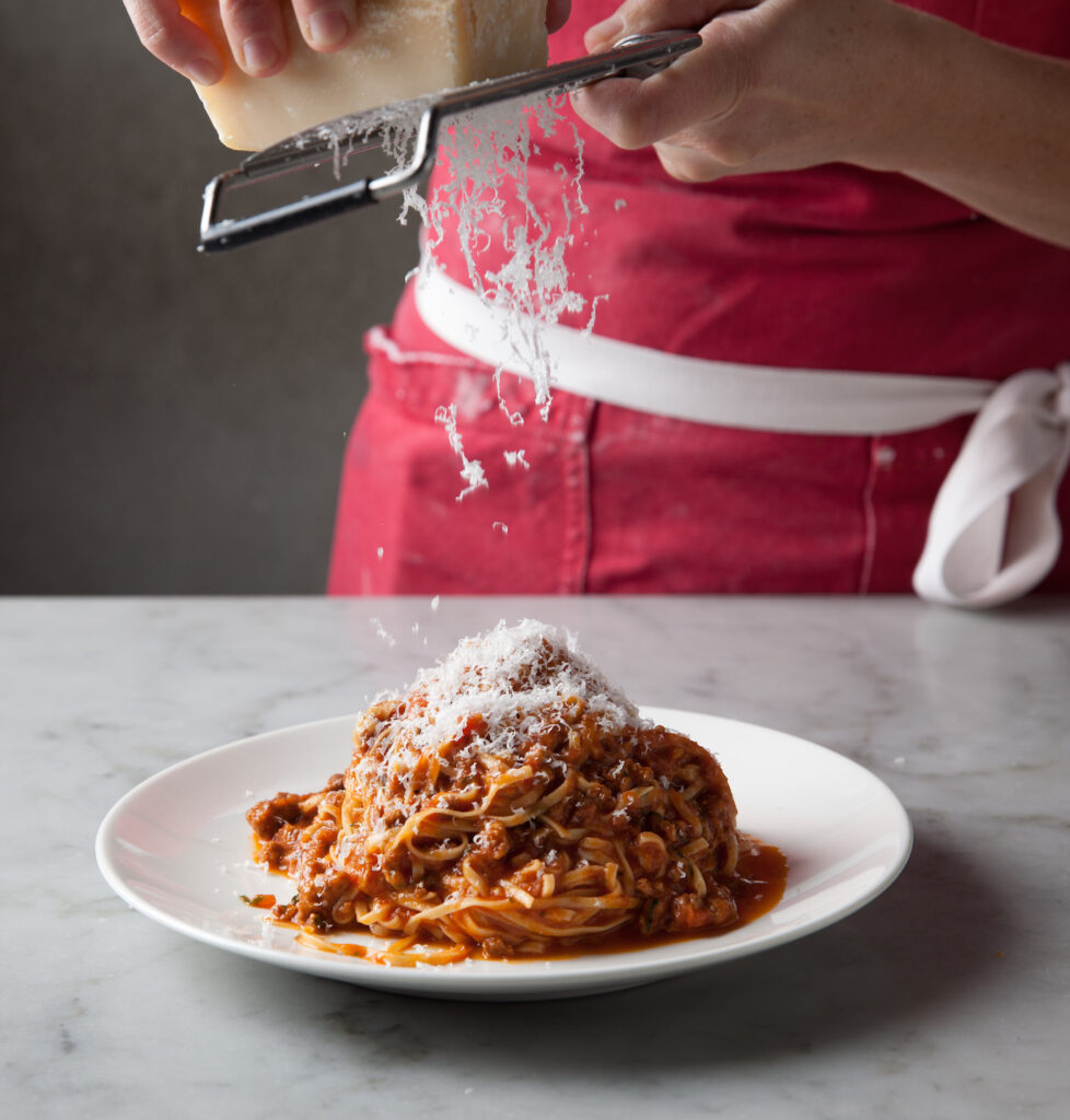 A person in an apron grates fresh parmesan onto a plate of pasta
